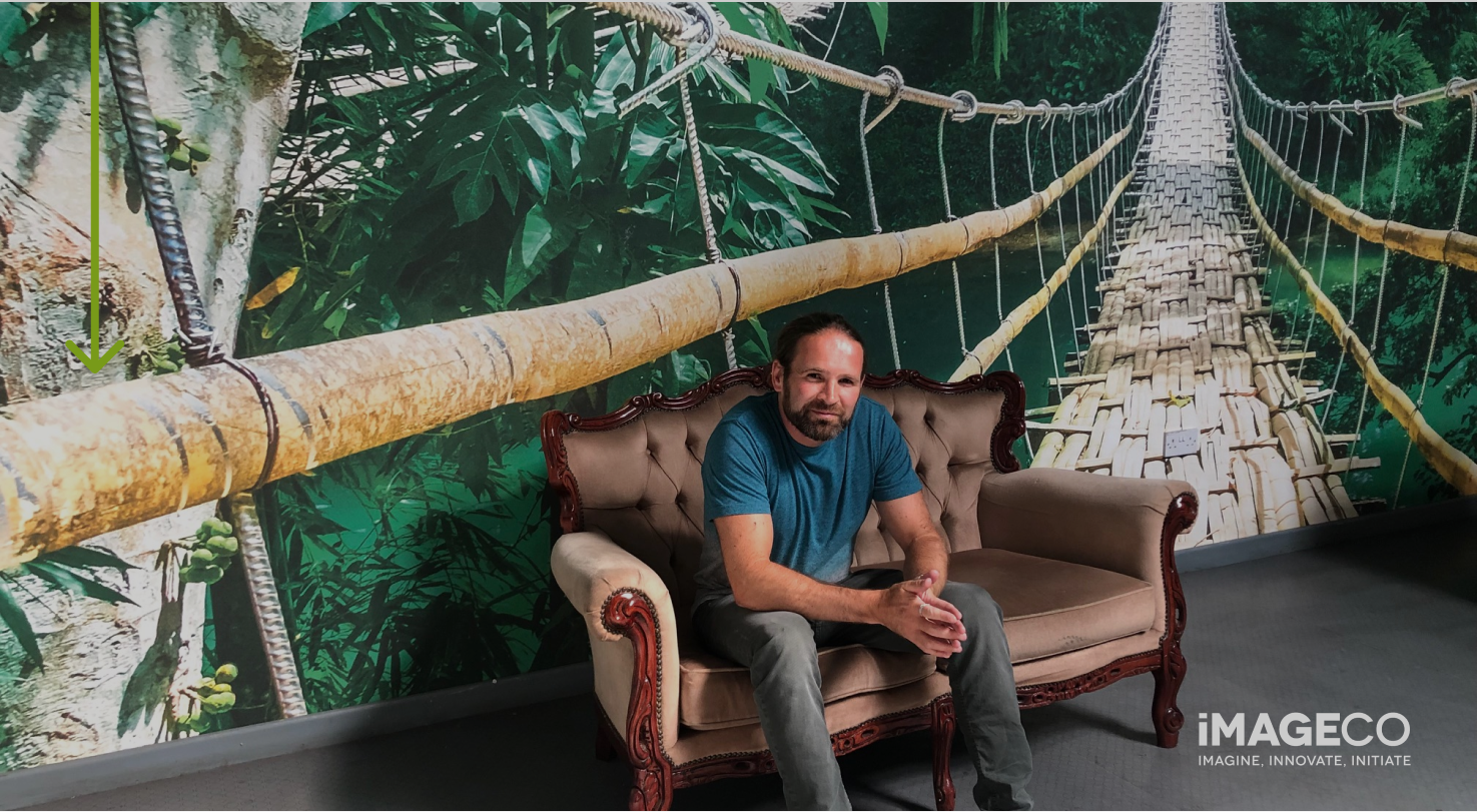 Imageco Managing Director, Nathan Swinson Bullough sits on a brown sofa in front of green bespoke wallpaper